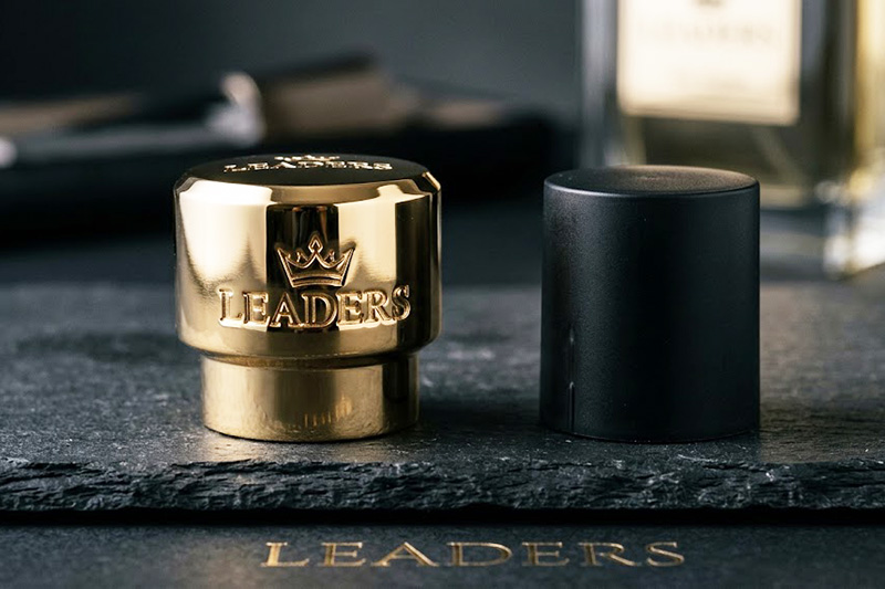 Gold die-cast Zamak perfume cap by Leaders compared next to a plain black plastic perfume cap on a dark slate surface.