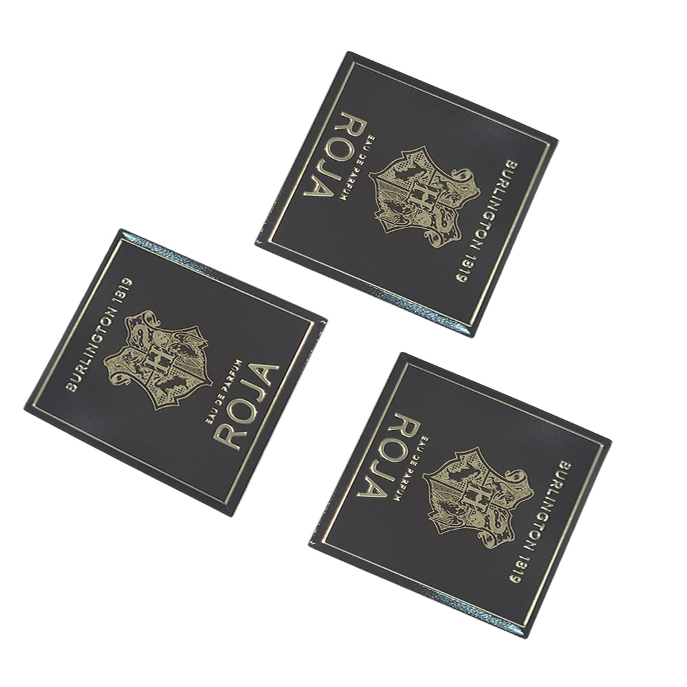 Three identical square black aluminum plaques with printed gold crests and lettering for perfume packaging.