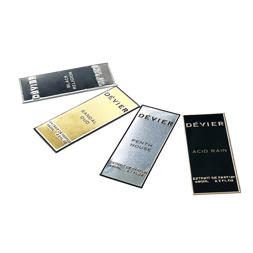 Fan-shaped arrangement of custom DÉVIER aluminum perfume labels displaying gold, silver, glossy black, and back-side view with adhesive tape.